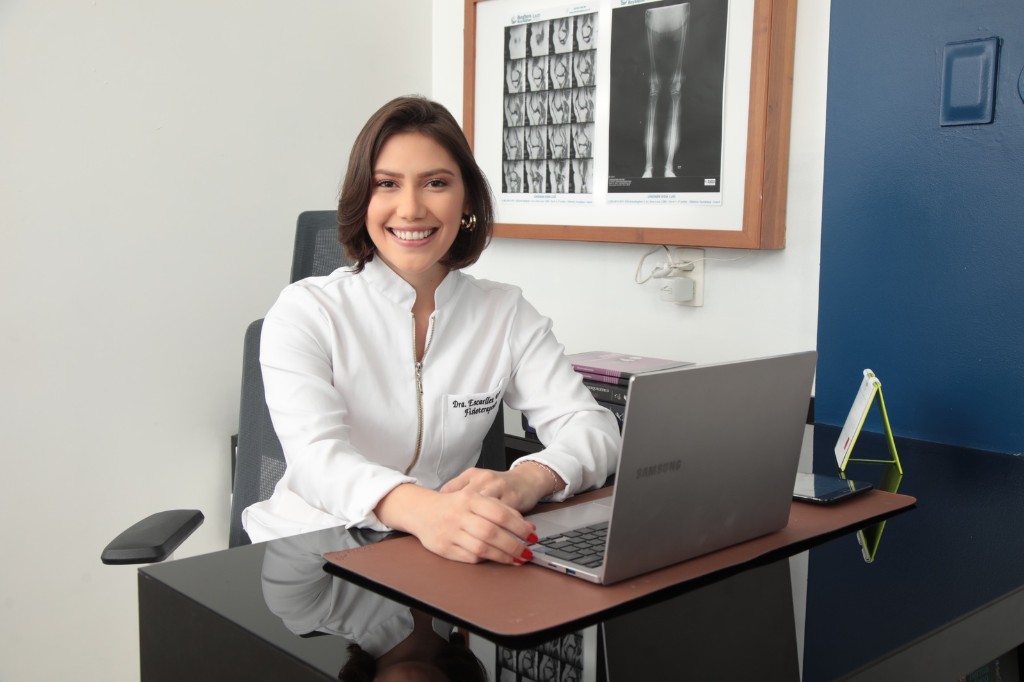 Dra. Escarllet Alves sentada à mesa do consultório, com radiografias de membros inferiores ao fundo