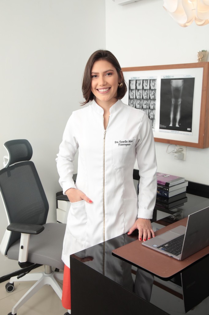 Dra. Escarllet Alves, fisioterapeuta, sorrindo em pé no consultório